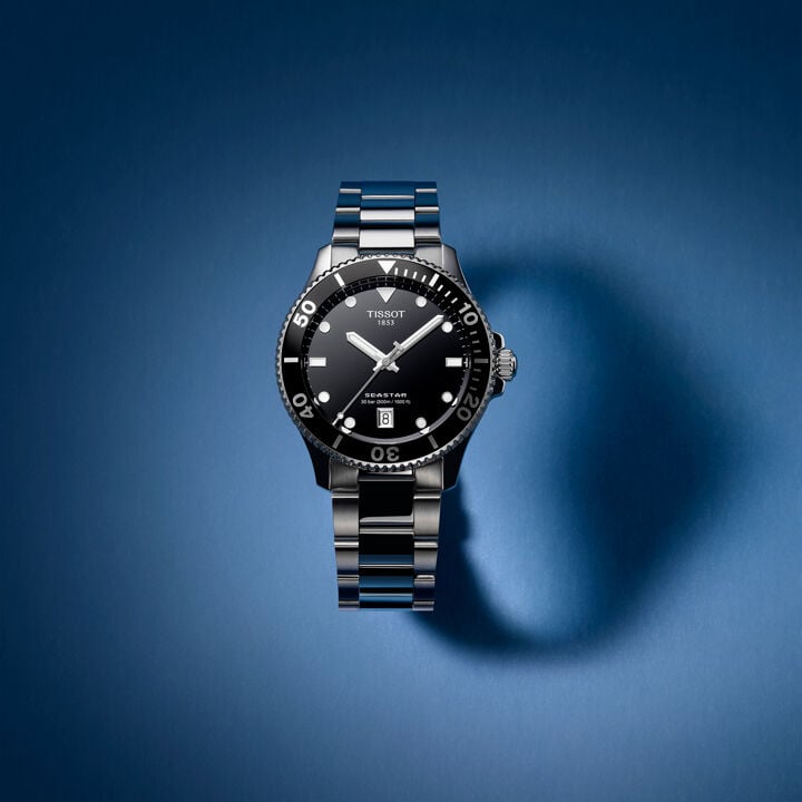 Tissot Seastar watch with a black dial, stainless steel bracelet, and unidirectional bezel, displayed against a gradient blue background