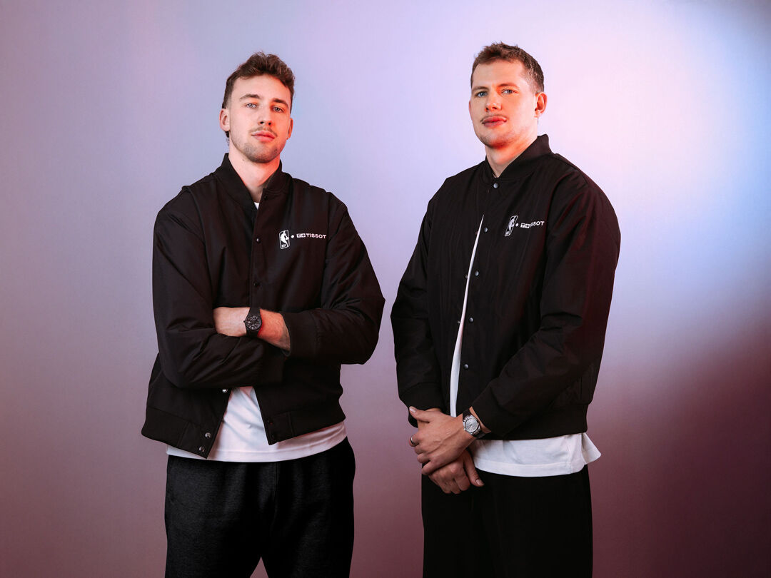 Moritz and Franz Wagner stand side by side wearing black Tissot-branded jackets, each wearing a Tissot watch, facing the camera with a confident, composed posture against a soft gradient background.