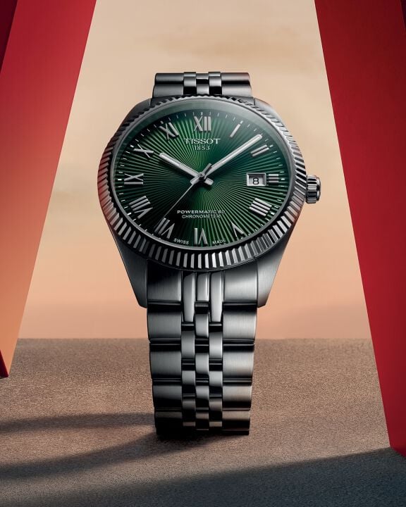 Relógio Tissot Ballade COSC com mostrador verde, ladeado por duas estruturas vermelhas nas laterais, diante de um pôr do sol em tons quentes ao fundo.