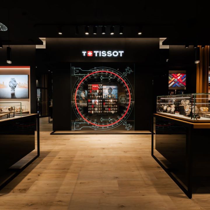 Front view of the Tissot boutique in Lausanne, showcasing a central digital wall display resembling a watch mechanism, with various watch models arranged inside. Illuminated glass cases on either side display a variety of wristwatches, under warm lighting and wooden flooring.