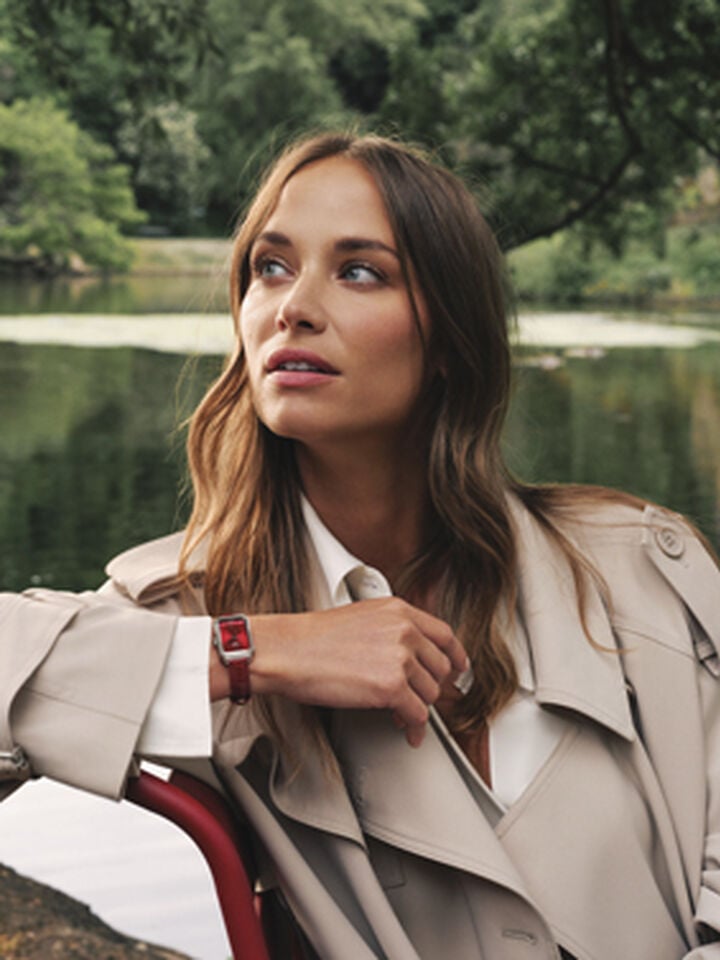 Agathe Teyssier, a friend of the brand, in a beige trench coat highlighting the Tissot SRV watch with a red leather strap, seated outdoors by a calm lake with greenery in the background.