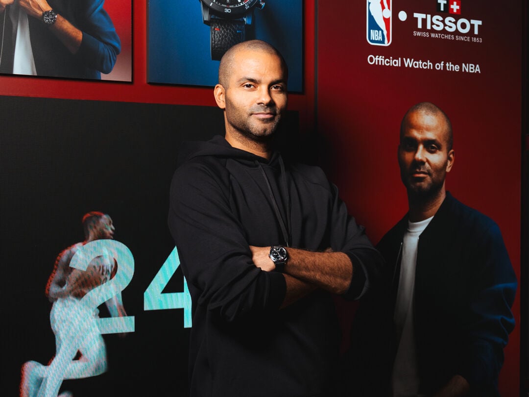  Tony Parker leans against a wall, posing in a three-quarter profile while holding a basketball in his hands. He wears the Tissot Supersport on his wrist, against a red-black gradient background.