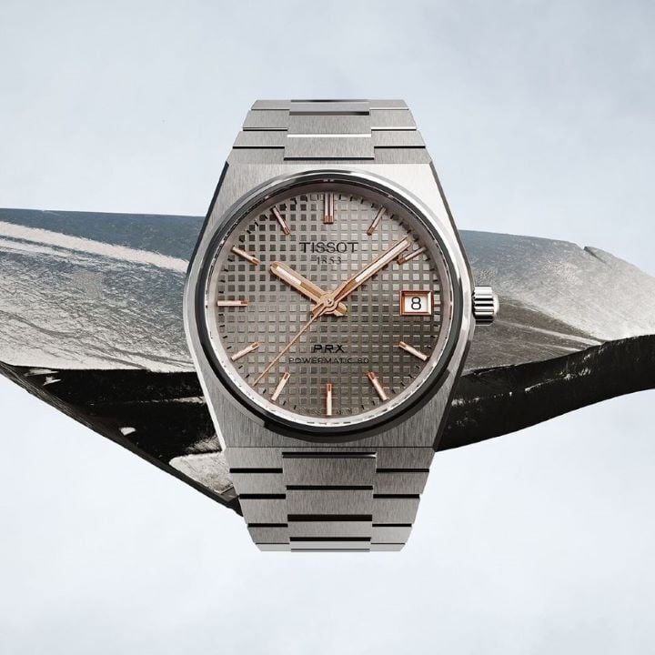 Relógio Tissot PRX 38mm Titan sobre um fundo cinza neutro com um pedaço de titânio ao fundo