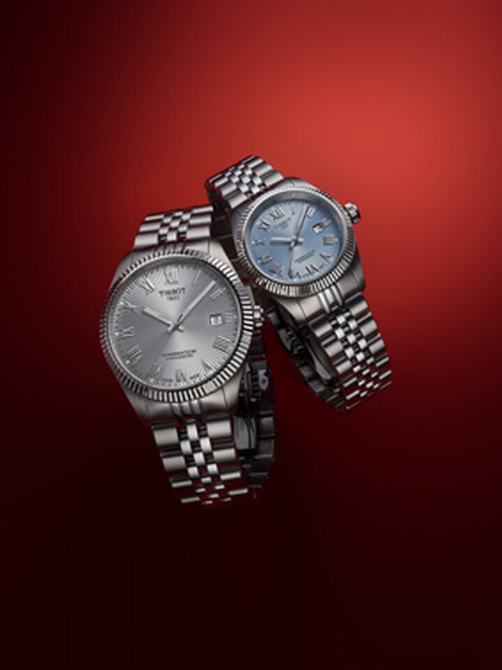 Two Tissot Ballade COSC watches with steel bracelets, silver and blue dials, presented as a duo on a gradient red background.