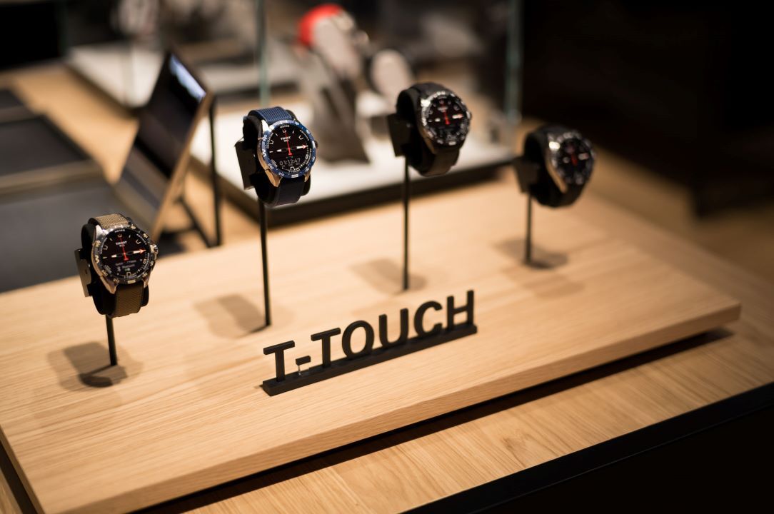 Nahaufnahme von Tissot T-Touch-Uhren auf einem hölzernen Stand, angeordnet mit eleganten schwarzen Haltern und einem Schild mit dem T-Touch-Logo im Vordergrund.
