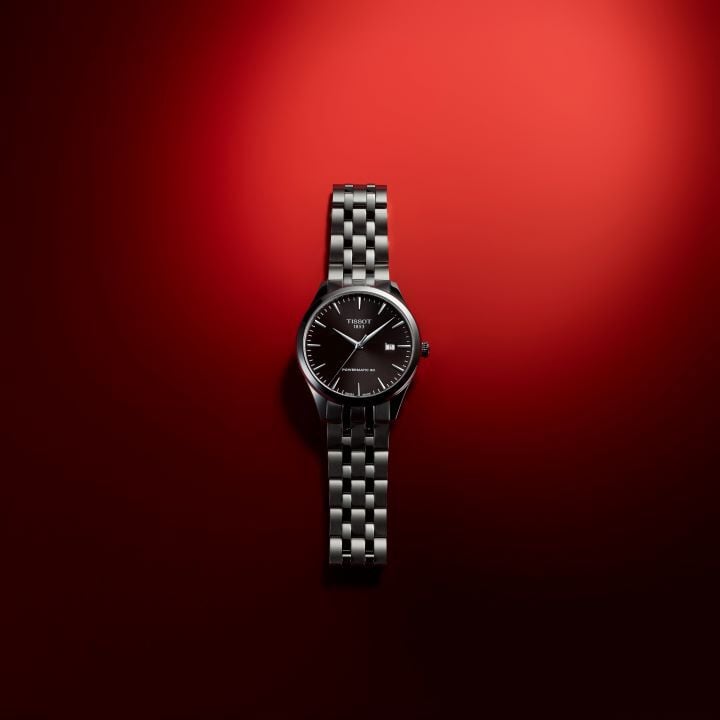 Relógio Tissot Classic Dream Powermatic 80 todo em aço, mostrador preto com índices prateados, caixa e bracelete em aço, sobre fundo vermelho.