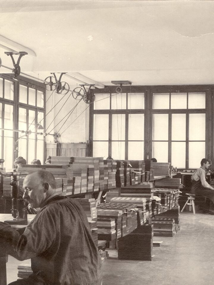 Werkplaats voor het afwerken van horloges van Tissot in Le Locle, begin 1900, met arbeiders die onderdelen in elkaar zetten en machines met riemaandrijving boven hun hoofd.