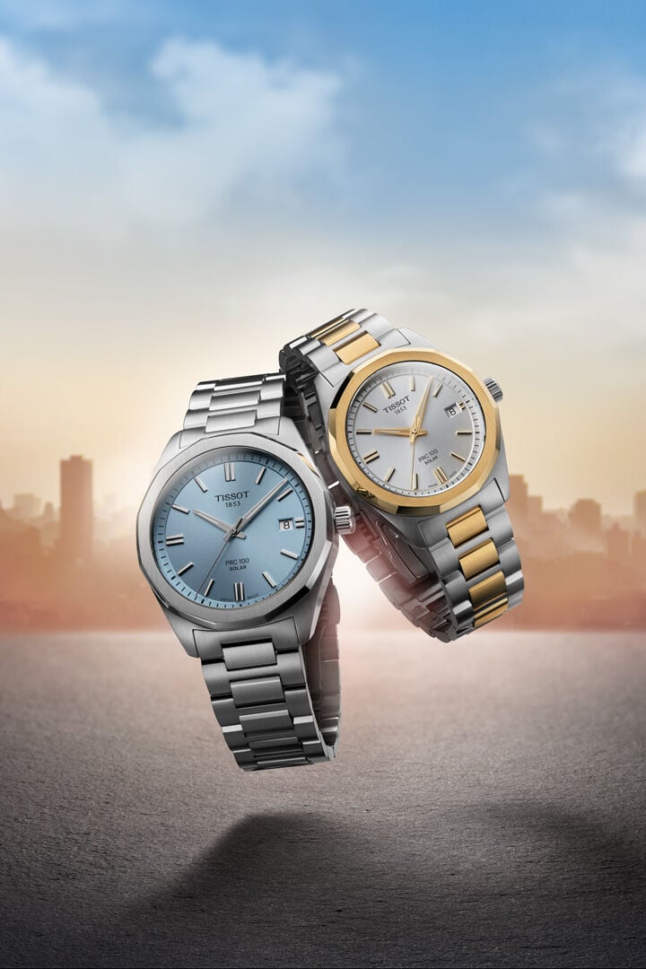 Zwei Tissot PRC 100 Solar Armbanduhren vor einer Skyline, &uuml;ber der die Sonne aufgeht.