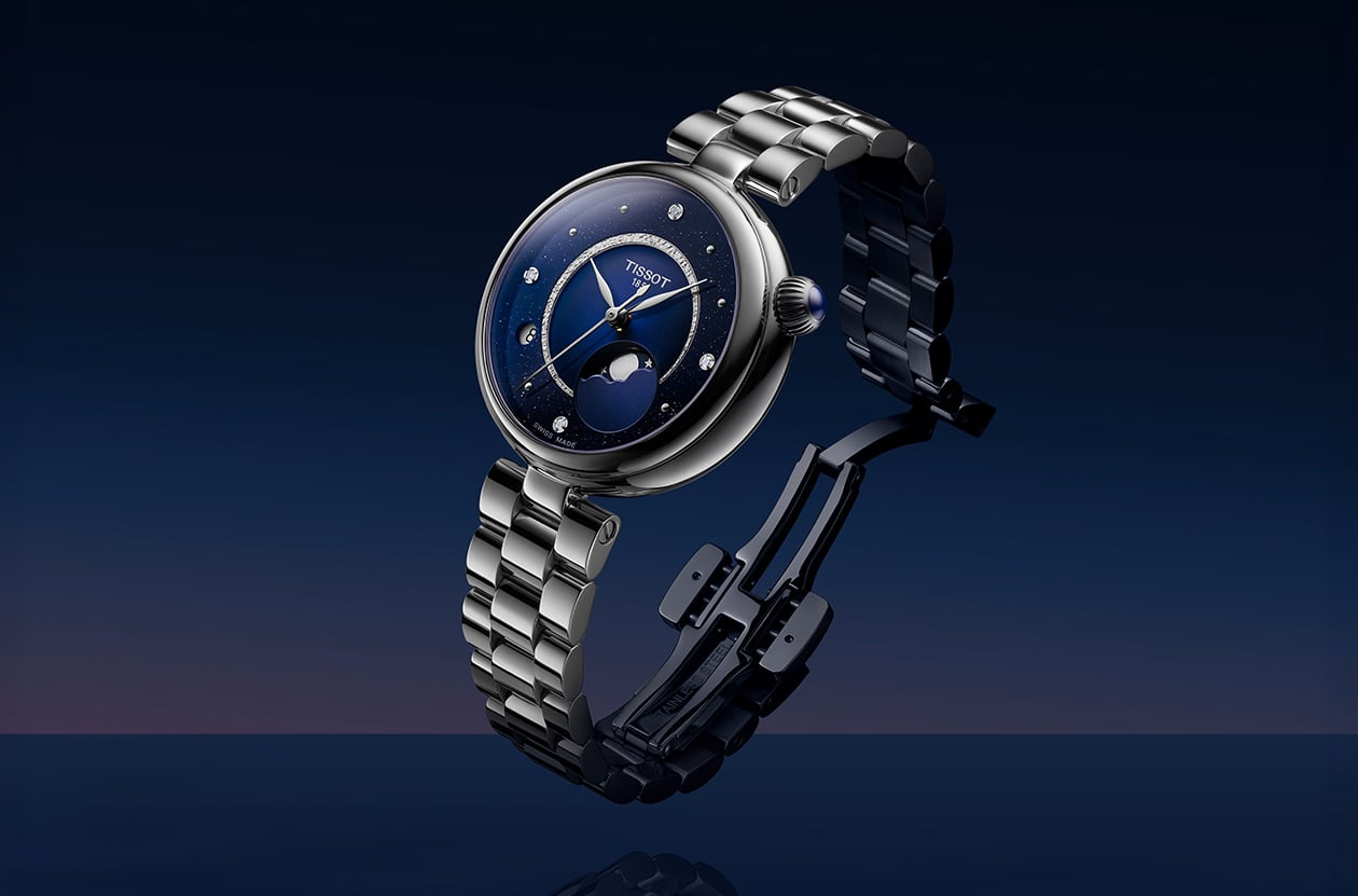 Tissot D&eacute;sir Moonphase Damenuhr mit dunkelblauem Zifferblatt, Diamanten, Mondphasenanzeige und Edelstahlarmband vor einem dunkelblauen Hintergrund.