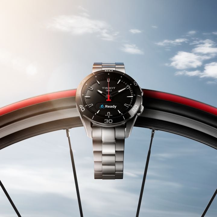 Tissot T-Touch Connect Sport watch suspended on a bicycle wheel, with a sturdy case, digital and analog display, and steel bracelet, against a blue sky background with a few clouds.