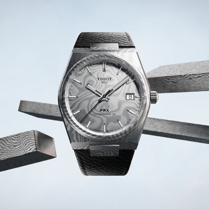 Tissot PRX 38mm Damascus watch on a light blue neutral background with pieces of Damascus steel in the background