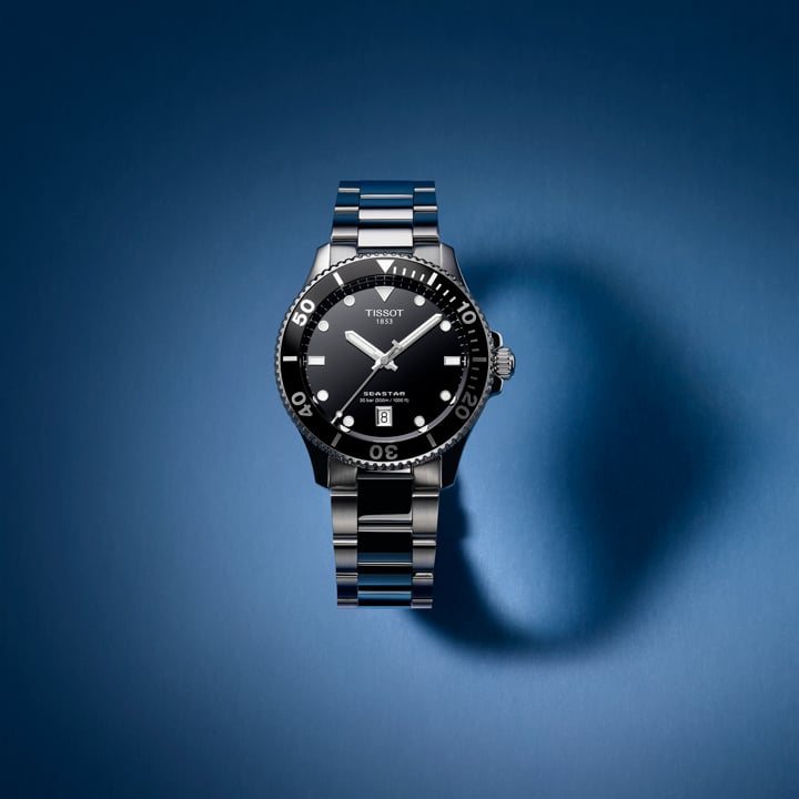 Tissot Seastar watch with a black dial, stainless steel bracelet, and unidirectional bezel, displayed against a gradient blue background