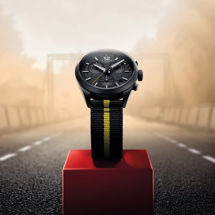 Tissot PR100 Tour de France chronograph watch with a black dial and black strap displayed on a cycling podium.
