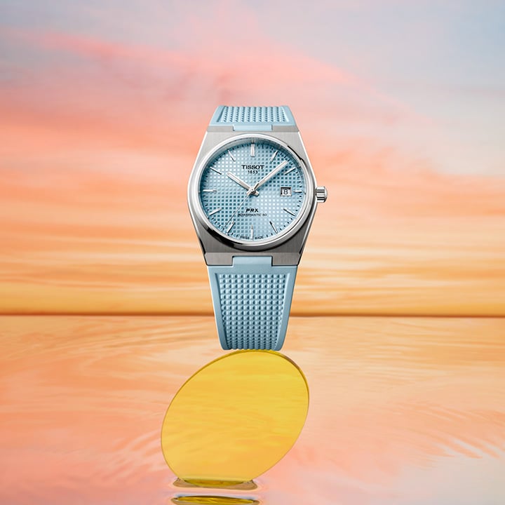 De PRX Light Blue wordt gepresenteerd boven een reflecterende achtergrond met de warme kleuren van een zonsondergang.