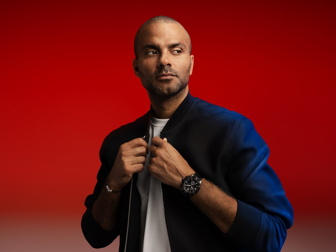  Tony Parker leans against a wall, posing in a three-quarter profile while holding a basketball in his hands. He wears the Tissot Supersport on his wrist, against a red-black gradient background.