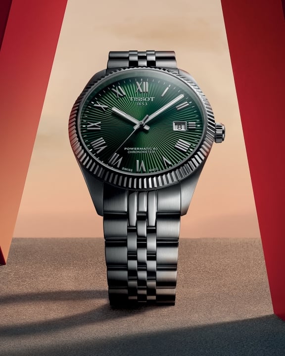 Tissot Ballade COSC Uhr mit gr&uuml;nem Zifferblatt, flankiert von zwei roten Strukturen an den Seiten, vor einem Sonnenuntergang in warmen Farbt&ouml;nen im Hintergrund.