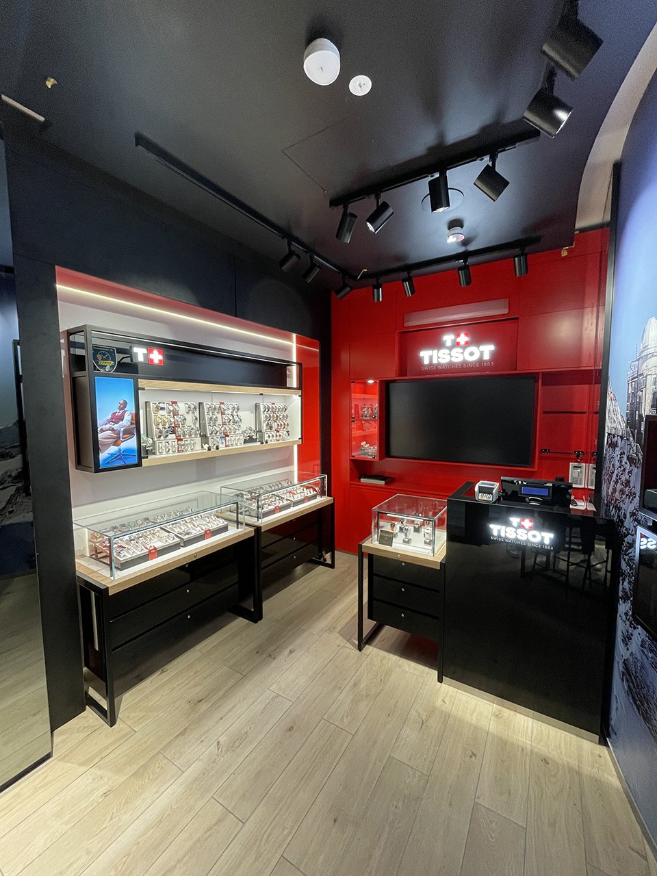 Dieses Bild zeigt eine moderne, kompakte Tissot-Boutique mit roter, schwarzer und wei&szlig;er Dekoration. Uhren sind in Glask&auml;sten ausgestellt, und das Tissot-Logo ist prominent an der Wand zu sehen. Der elegante Raum kombiniert klare Linien mit einem hellen Holzboden als Kontrast.