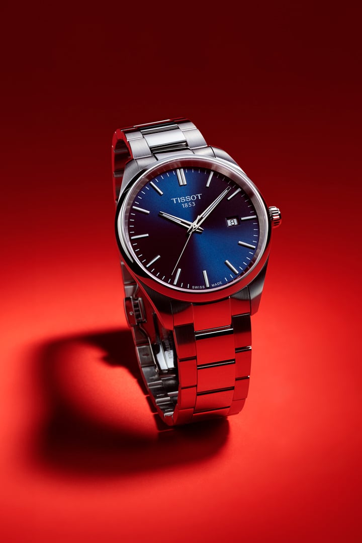 Tissot PR 100 Armbanduhr mit blauem Zifferblatt und Edelstahlarmband vor dunkelrotem Hintergrund.