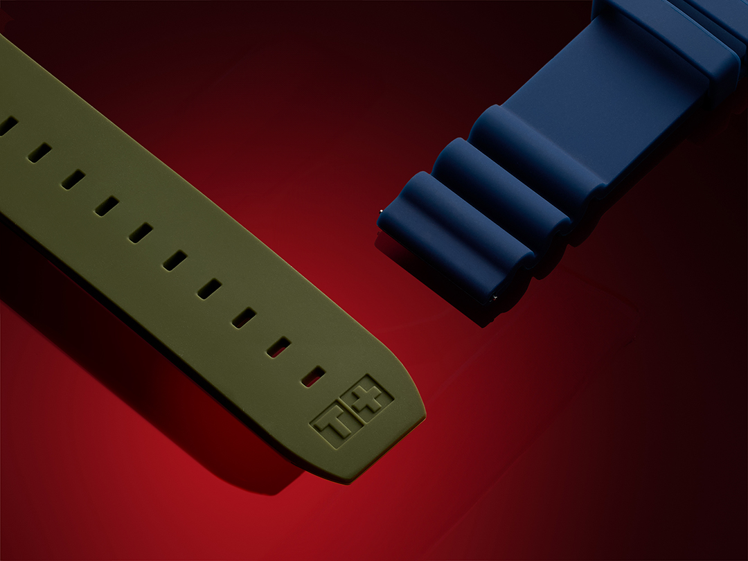 Close-up view of two Tissot rubber watch straps: one in olive green with a smooth finish and embossed 'T+' logo, and the other in dark blue with a distinctive wave pattern, set against a deep red background