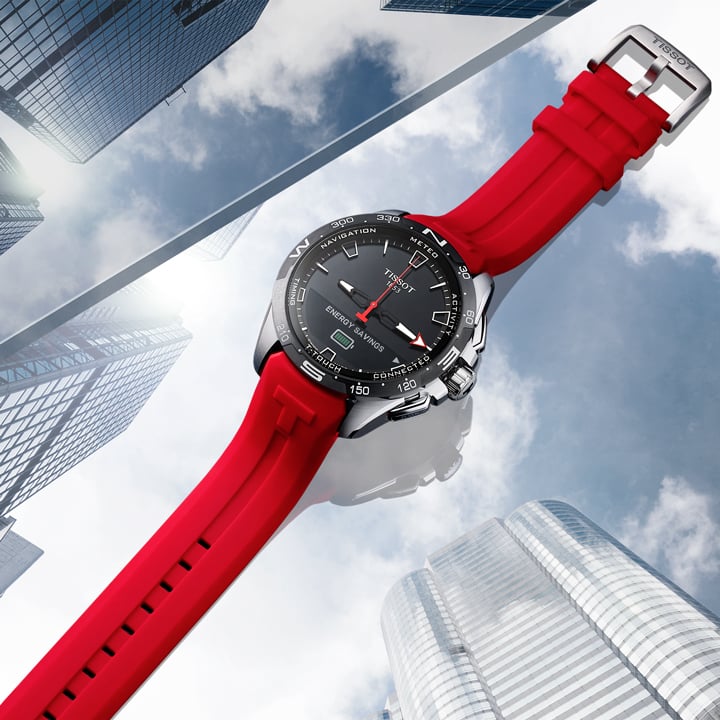 Tissot T-Touch Connect Solar smartwatch with a black dial, stainless steel case, and red silicone strap against a city skyline.