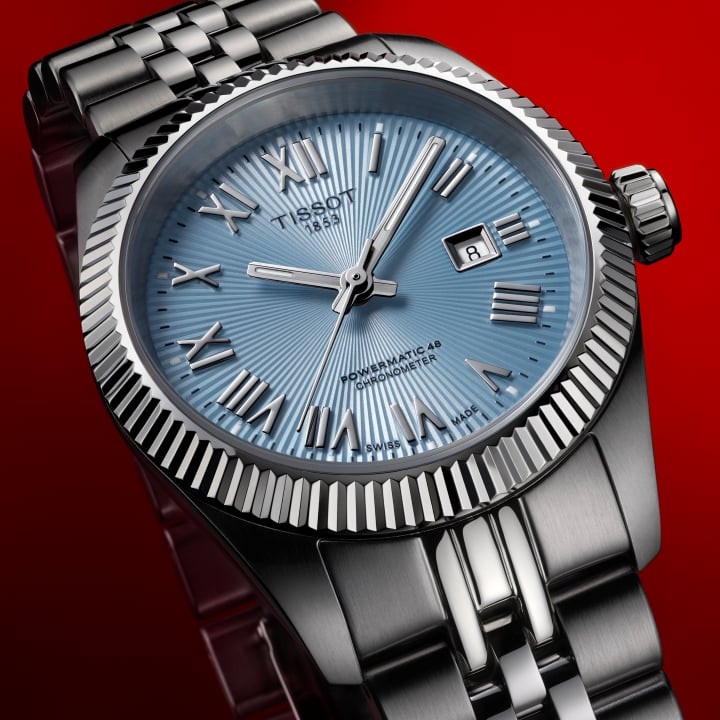 Tissot Ballade COSC Uhr mit Stahlarmband und blauem Zifferblatt, pr&auml;sentiert auf einem roten Farbverlauf-Hintergrund.