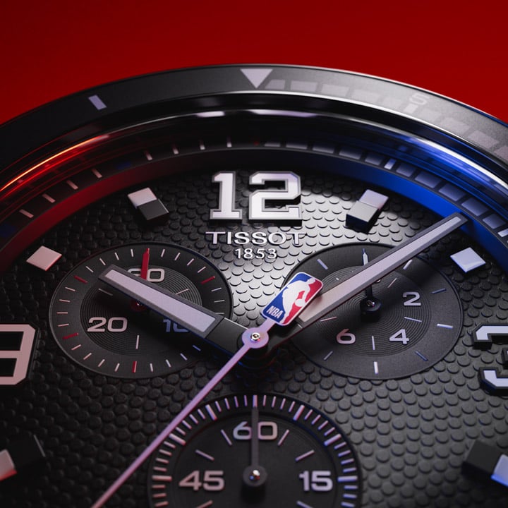 Extreme close-up of the Tissot Supersport Chrono NBA Edition watch dial, highlighting the textured black surface, silver numerals, chronograph sub-dials, and the NBA logo on the second hand. The bezel features precise markings, with red and blue lighting reflections enhancing the sporty and dynamic design.