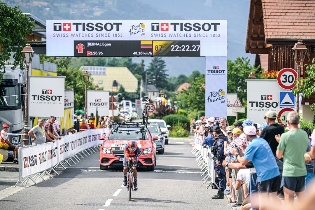 A cyclist crosses the finish line at a Tour de France stage, with a large Tissot-branded timer displaying the rider's timing and name, Egan Bernal, along with his speed.
