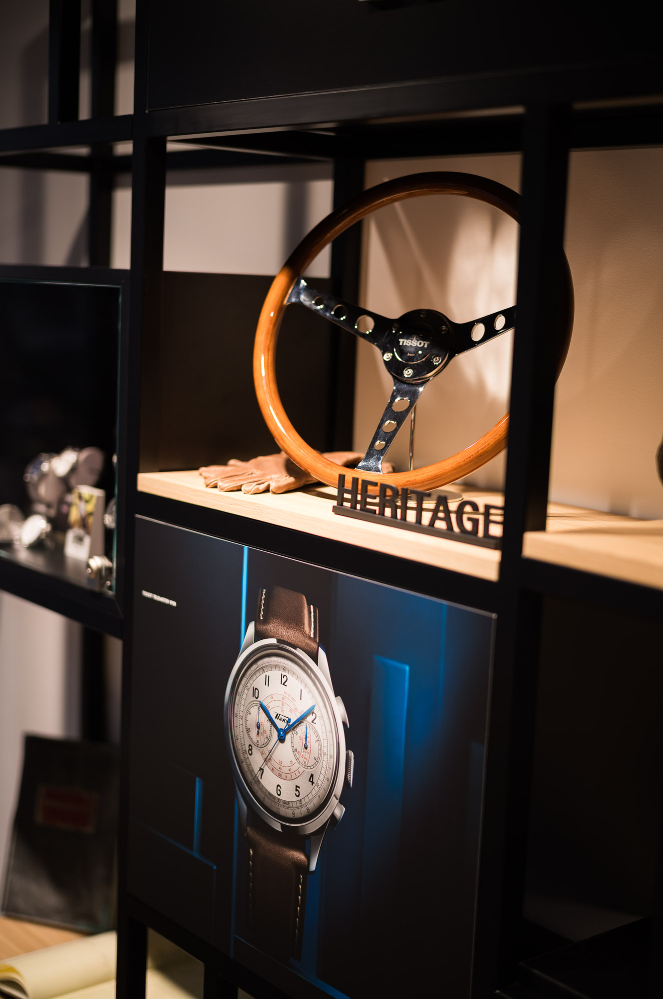 Ein h&ouml;lzernes Lenkrad und Vintage-Handschuhe, die auf einem Regal neben einem Uhrenposter in einer Glaskabine ausgestellt sind.