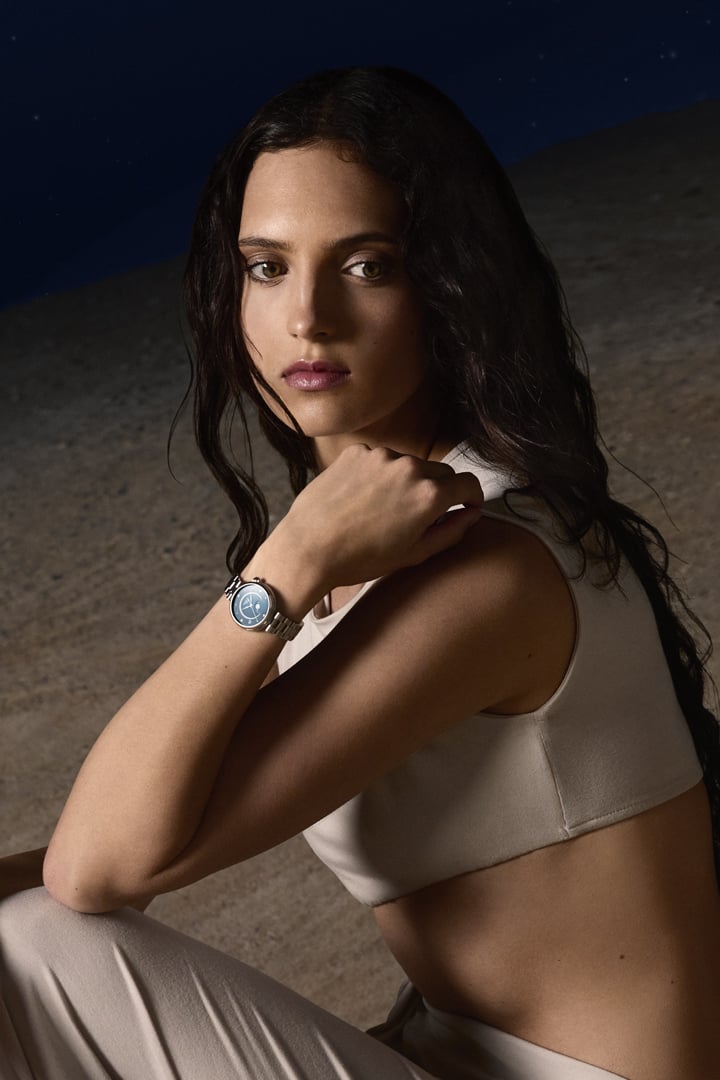 Woman seated on a moonlike landscape at night, wearing a D&eacute;sir Moonphase watch with a blue dial beneath a starry sky.