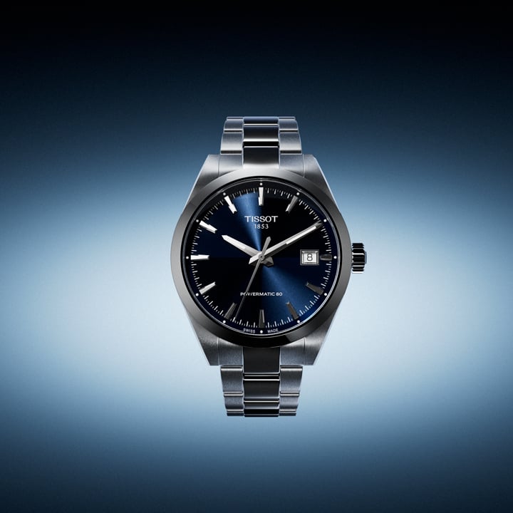 Eine Tissot Gentleman 38mm Herren-Armbanduhr mit dunkelblauem Zifferblatt und Edelstahlarmband vor einem bl&auml;ulichen Hintergrund.