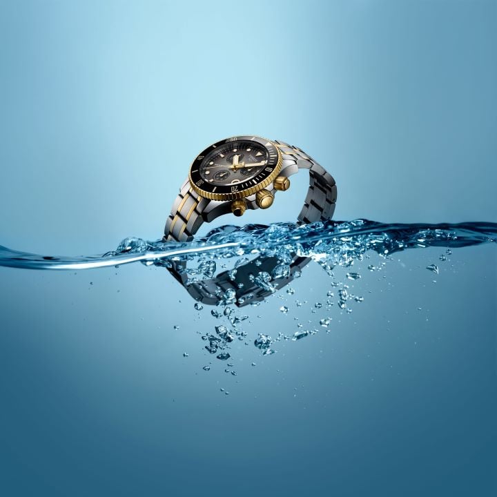 Tissot Seastar 38mm is partially immersed in water, highlighting its two-tone steel and gold case, black dial with rotating bezel, and gold push buttons, against a light blue background evoking water resistance.