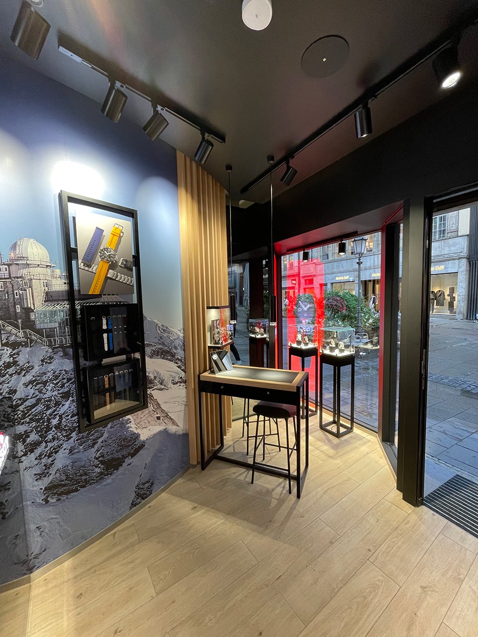 Das Interieur der M&uuml;nchner Boutique von Tisso. Auf der linken Seite zeigt ein gerahmtes Display eine Uhr mit bunten Armb&auml;ndern vor einer verschneiten Bergkulisse. 