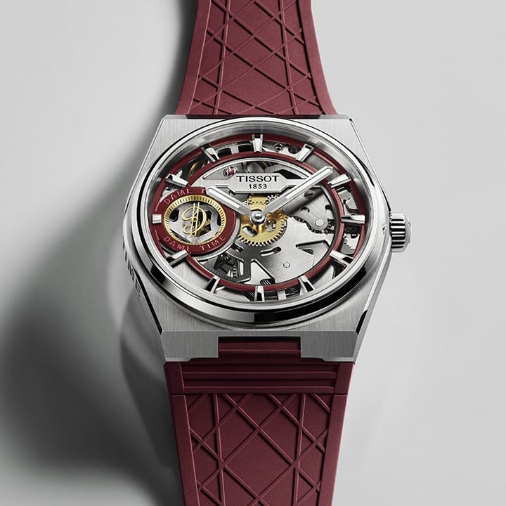 Tissot watch with a skeleton dial, silver-tone case, and burgundy rubber strap with a geometric pattern against a gray background.