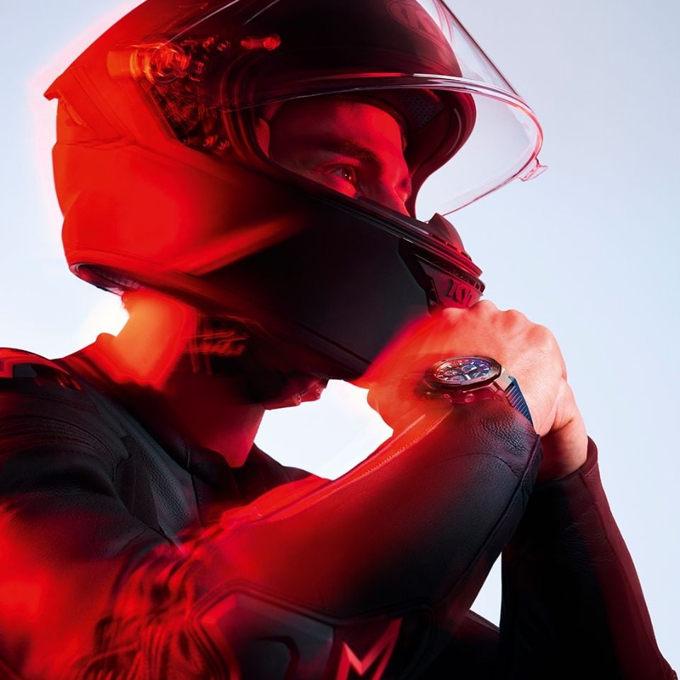 Na lekko rozmazanym czerwonym tle obraz ukazuje Enea Bastianiniego w kasku i stroju motocyklowym, z zegarkiem Tissot T-Race MotoGP Chronograph 2024 Limited Edition na nadgarstku.