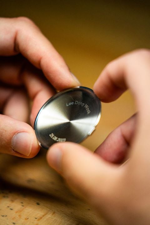 Close-up of a watch case back being held, engraved with "Lee Dong Wook" and a date "23.09.2023".