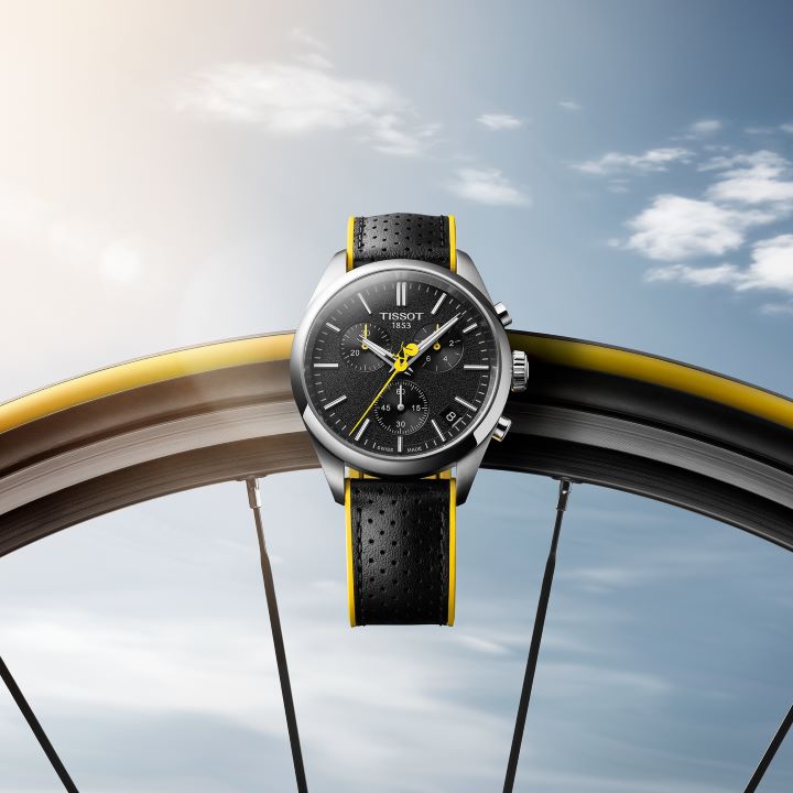 Tissot PR 100 Tour de France watch hanging on a bicycle wheel, featuring a black dial with yellow details and a black leather strap with yellow edges, set against a blue sky with scattered clouds.