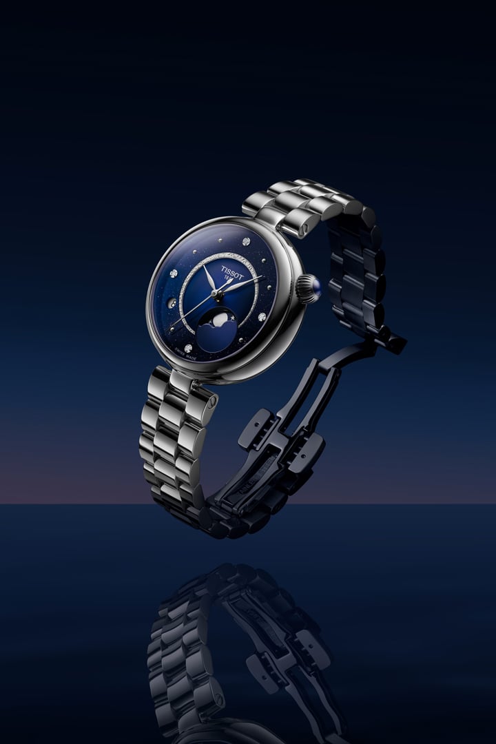 Tissot D&eacute;sir Moonphase Damenuhr mit dunkelblauem Zifferblatt, Diamanten, Mondphasenanzeige und Edelstahlarmband vor einem dunkelblauen Hintergrund.