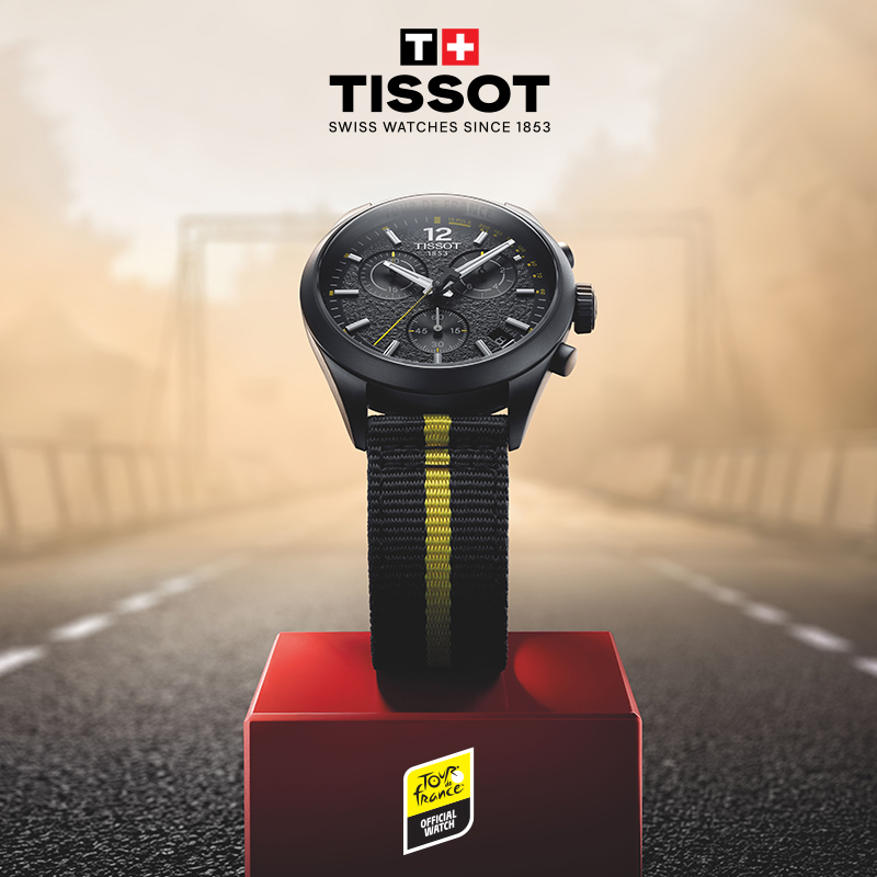 Black Tissot chronograph watch with yellow accents and a black fabric strap featuring a yellow center stripe, displayed on a red podium against a blurred road background
